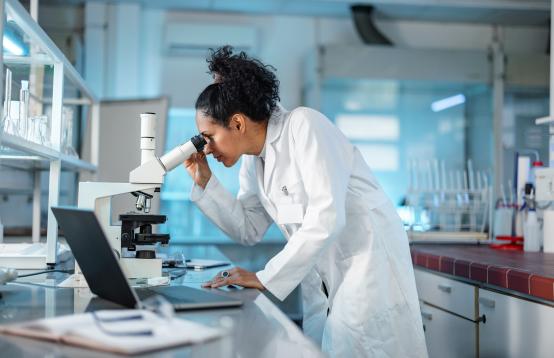 Woman in a lab coat with dark hair in a pony tail looking into a microscope in a laboratory.