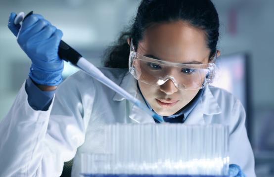 Female scientist with long dark hair in a pony tail wearing a white lab coat and safety glasses conducting an experiment.