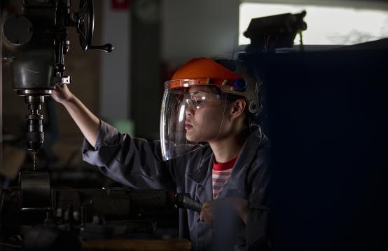 A person wearing a safety face mask dressed in overalls and working on a table top drill.