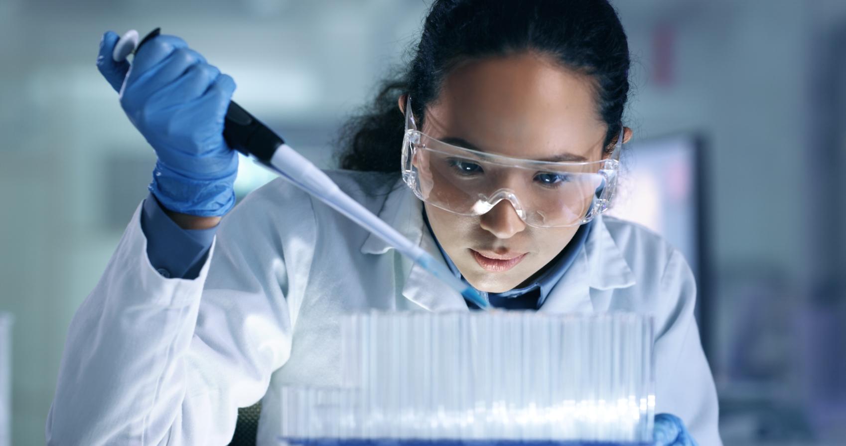 Female scientist with long dark hair in a pony tail wearing a white lab coat and safety glasses conducting an experiment.