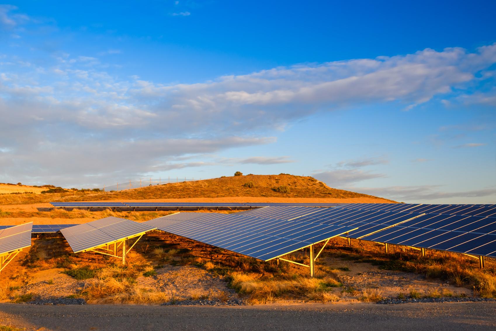 Landscape image of sand dunes with many solar panels and a blue sky with a few wispy clouds.