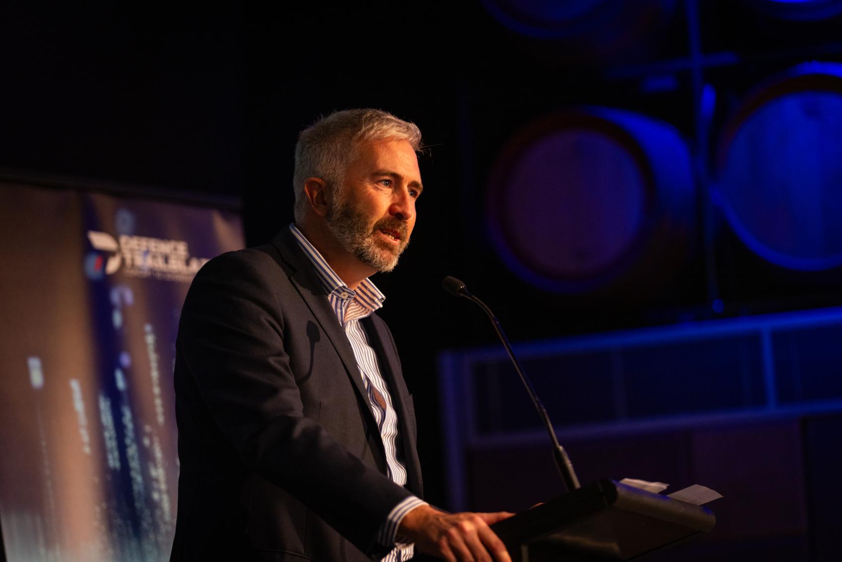 Image of a man with short grey hair and a beard, wearing a dark suit jacket with white business shirt speaking at a podium at a function.