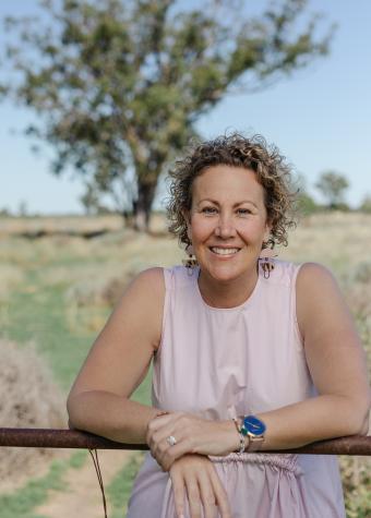 Photo from the waist up of a woman smiling, leaning on a paddock gate in the bush, wearing a sleeveless pink shirt, with short blonde curly hair smiling.