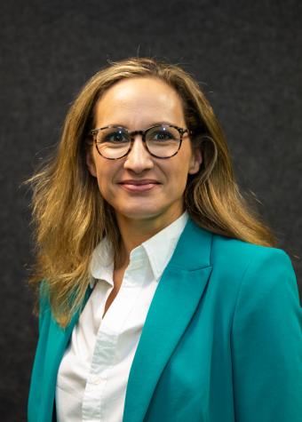 Upper body photo of a woman smiling, wearing a white collared, open necked shirt and a, aqua, long-sleeved business jacket.  She has long light brown hair and is wearing spectacles.