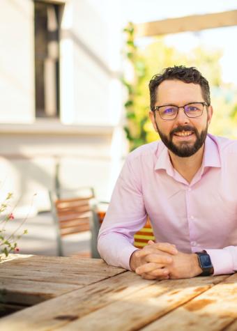 Photo of a man sitting at a wooden table in a sunny room.  He has short, dark hair and a beard and is wearing spectacles and a long sleeved, open necked business shirt with his hands clasped on the table.