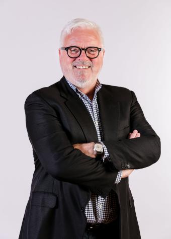 Photo of a smiling man wearing a dark business suit and light coloured, open necked business shirt, with short grey hair and beard wearing dark framed spectacles.