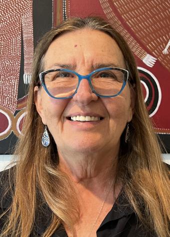 Head shot of a smiling lady with light brown long hair wearing light grey framed spectacles, dangling earrings and a black collared shirt. 